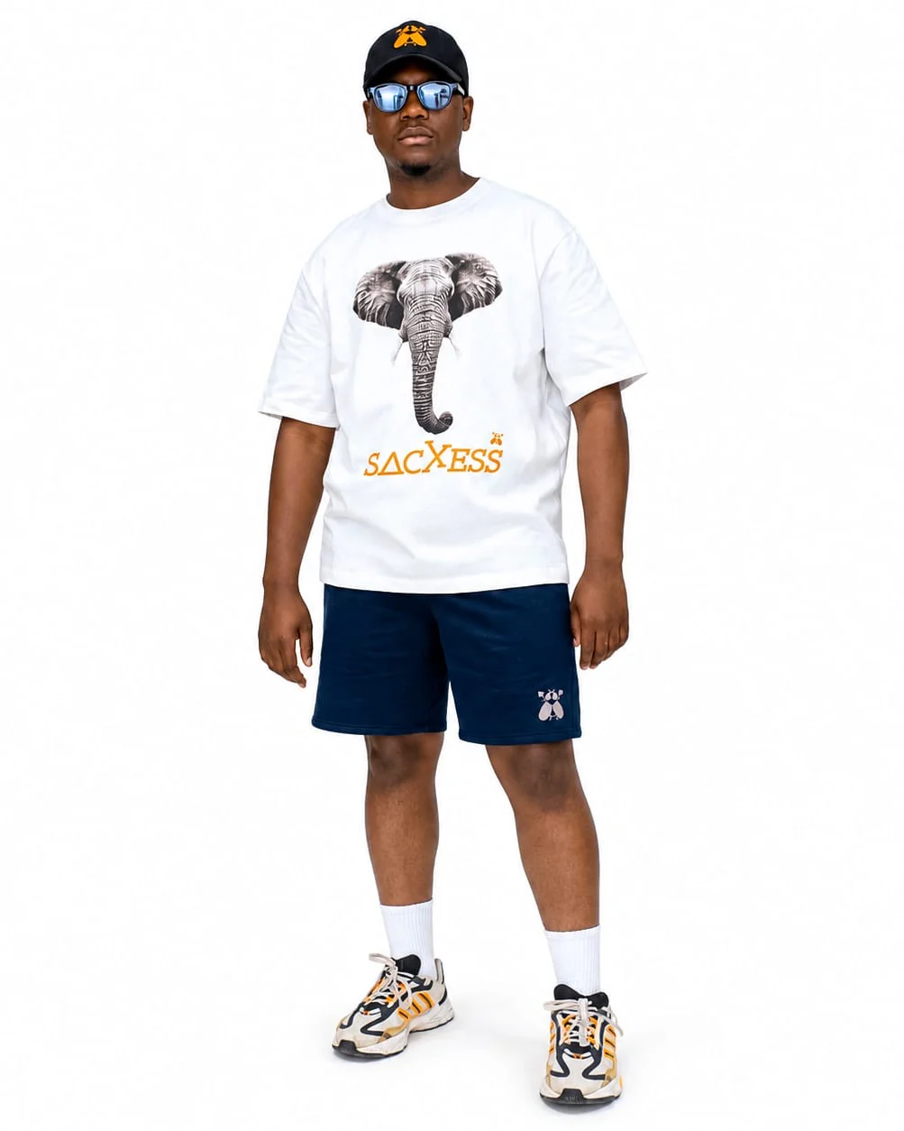 A model wearing the SACXESS Elephant Ed. Boxy Tee in Light with navy SACXESS shorts and a black SACXESS cap.